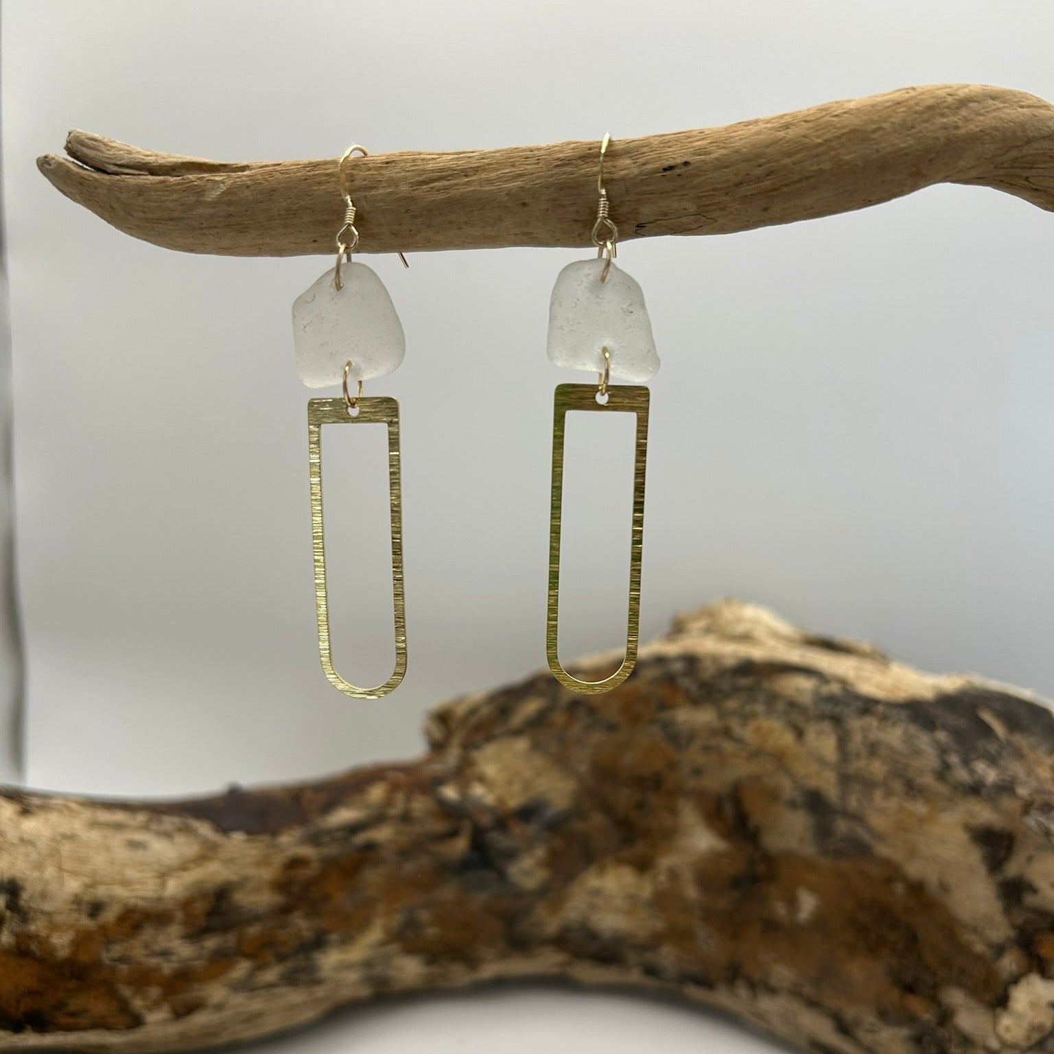 Joy Sea Glass earrings with brass D shape attachment.  
A pair of earrings made from clear sea glass and brass attachment.  Displayed on driftwood with a white backdrop.