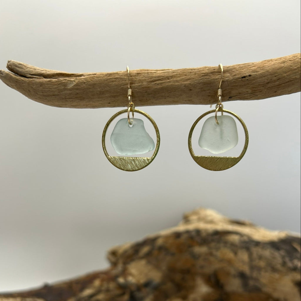 
Pair of earrings with clear sea glass and brass surround displayed on a piece of driftwood against a white background